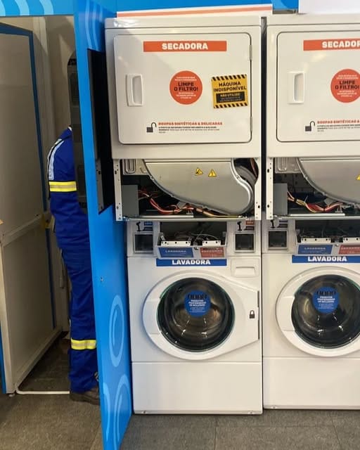 Técnico de uniforme azul fazendo manutenção em secadoras empilhadas sobre lavadoras numa lavanderia self-service