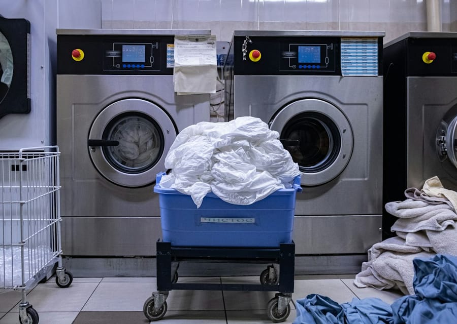 Lavadoras industriais de inox em uma lavanderia, com um carrinho de roupas brancas à frente